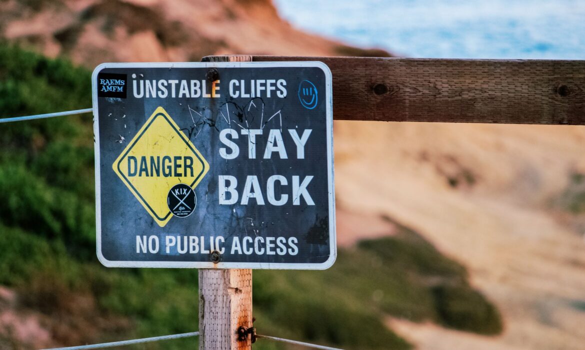 Warning sign indicating danger due to unstable cliffs, advises staying back. Outdoors setting.